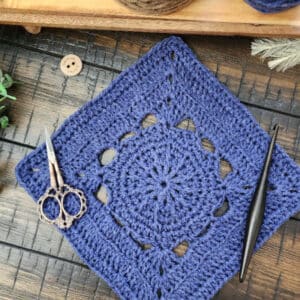 A textured 9-inch crochet square, the Fritzella Square, displayed on a wooden tray with greenery, showcasing its intricate stitch design.